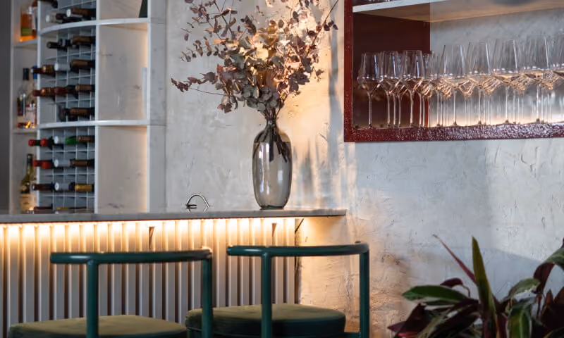 Modern bar counter with green stools, a vase with dried flowers, wine bottles, and hanging wine glasses.