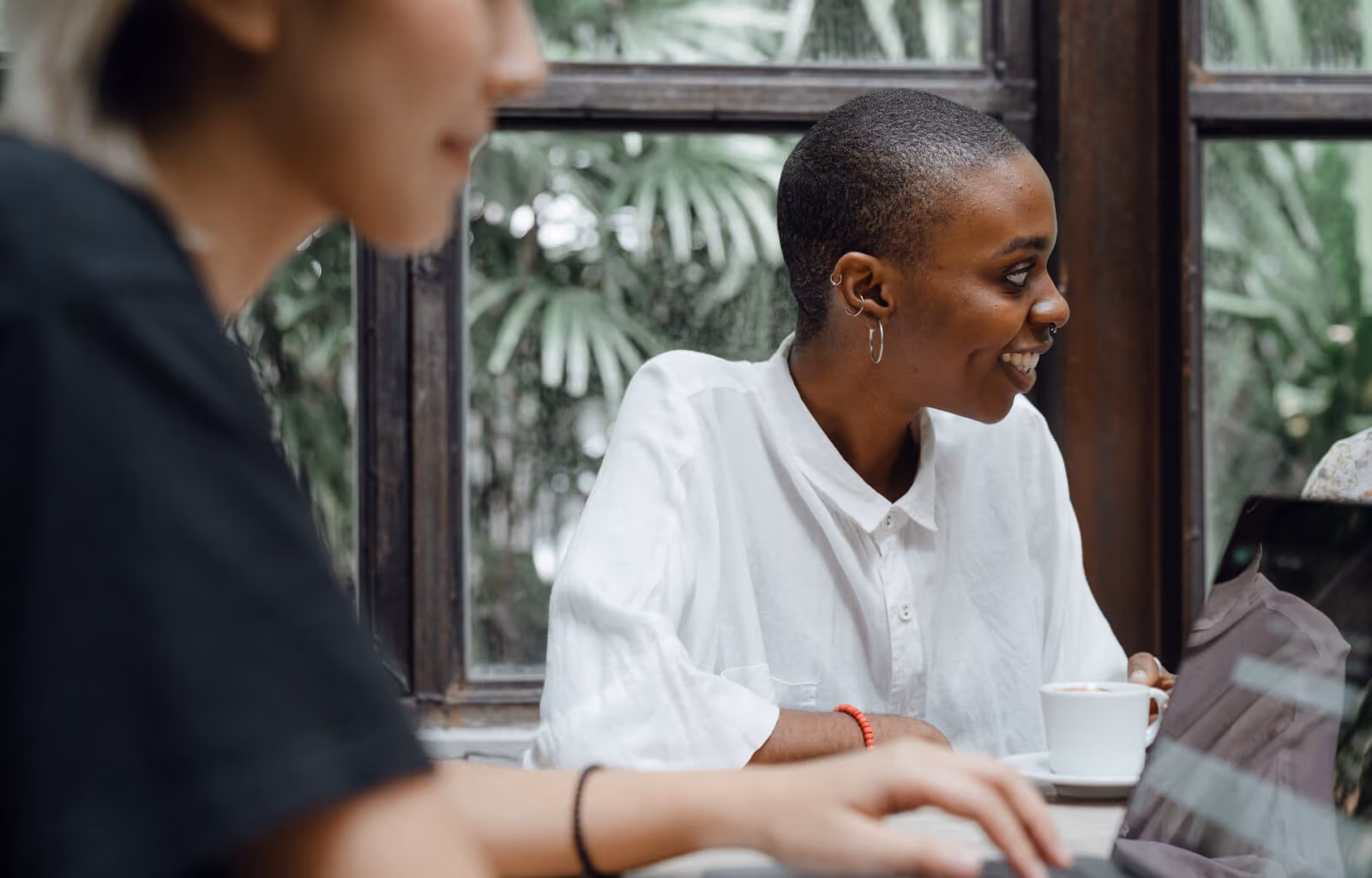 Person in white shirt smiling and holding a coffee cup near a window while another uses a laptop.