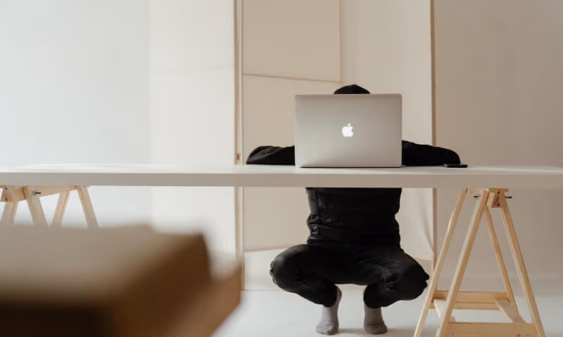 Person crouching under a white table with a laptop open on it in a minimal white room.