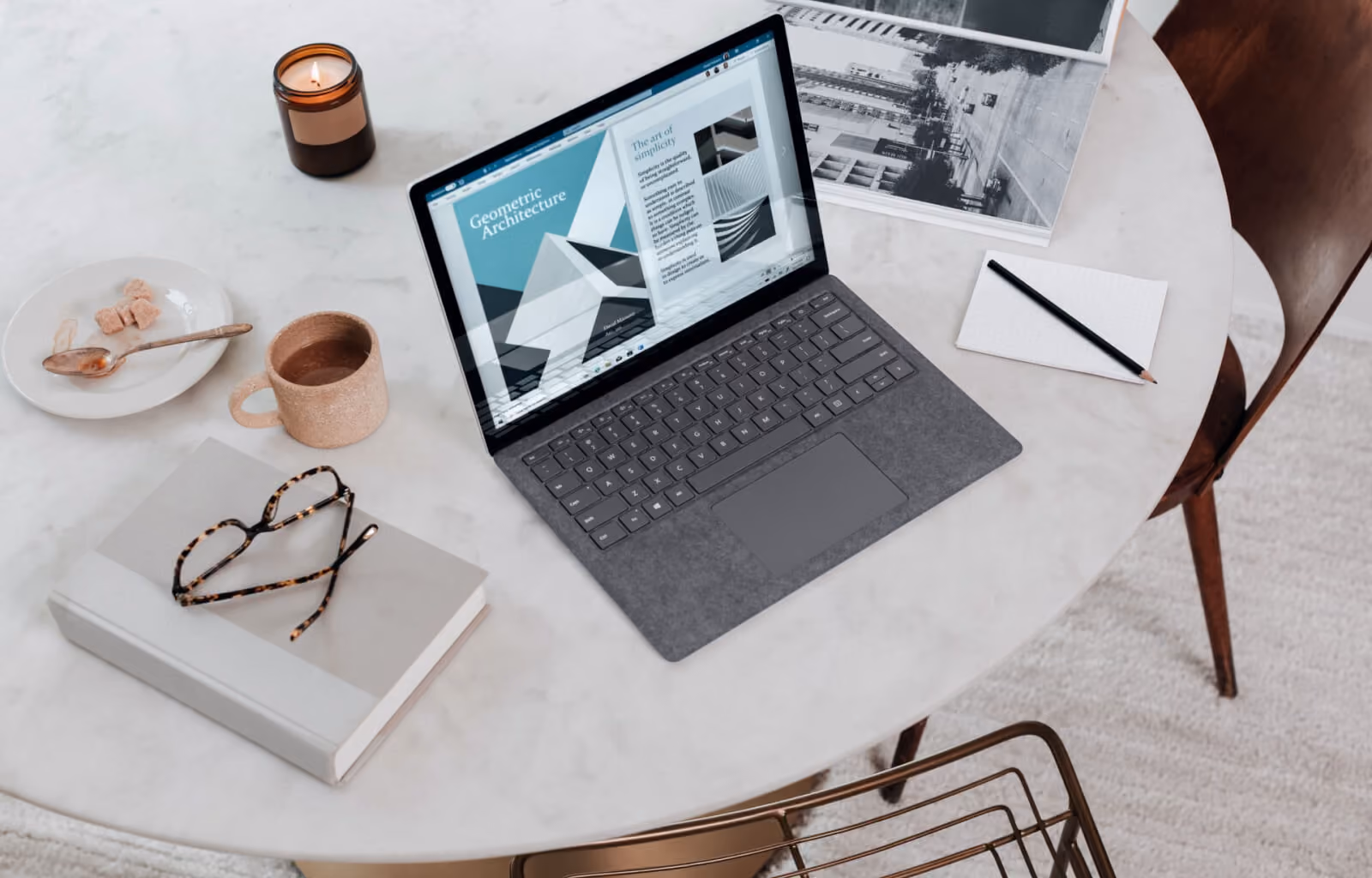 Marble table with laptop showing geometric architecture, mug, glasses on book, candle, and notepad with pencil.