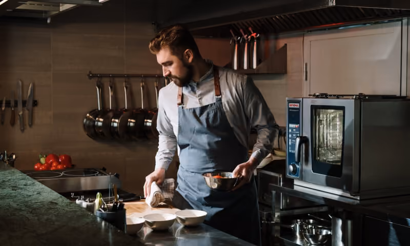 Chef in apron wiping counter with towel and holding bowl of vegetables in a professional kitchen.