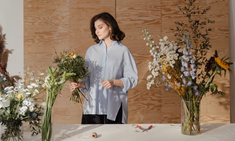 Woman arranging a bouquet of wildflowers at a table with various flowers and vase.