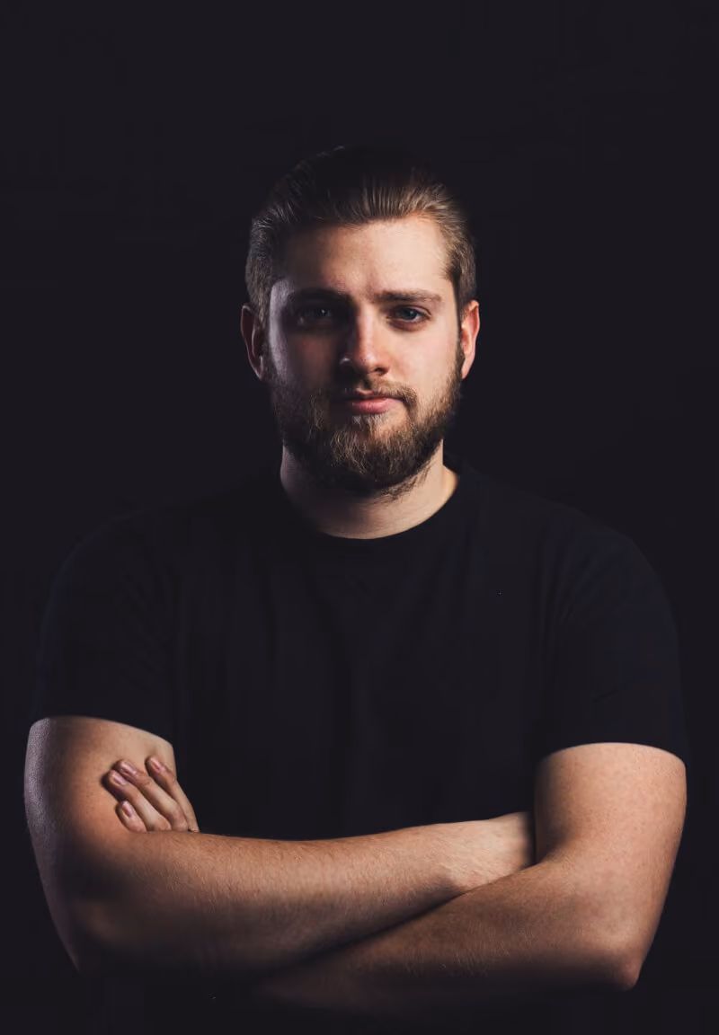 Bearded man in black shirt with arms crossed against dark background.
