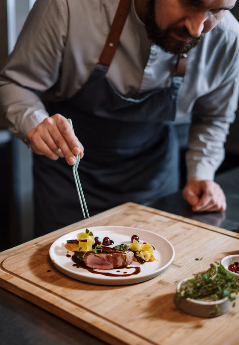 Chef wearing apron carefully garnishes plated gourmet dish with tweezers on wooden board.