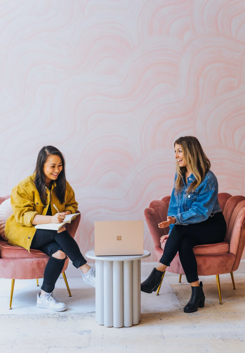 Two women sitting on pink chairs having a conversation with a laptop on a white round table between them.