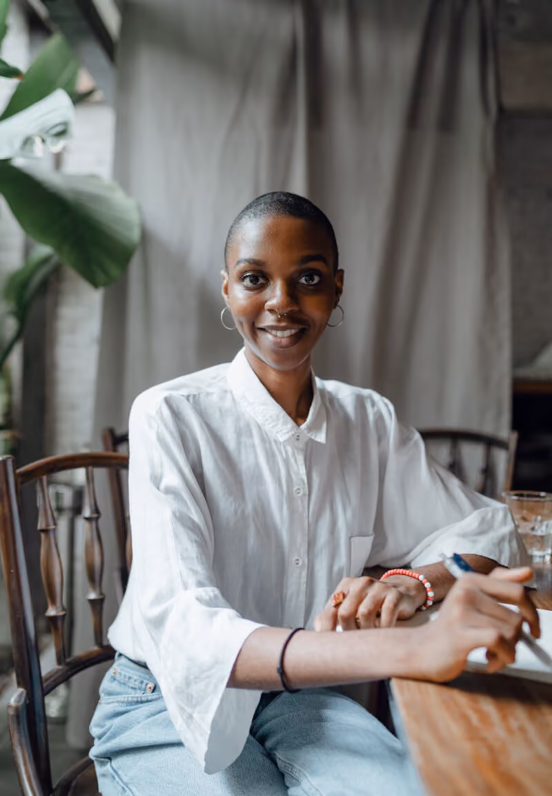 Smiling person with short hair wearing a white shirt sitting at a wooden table with a pen in hand.