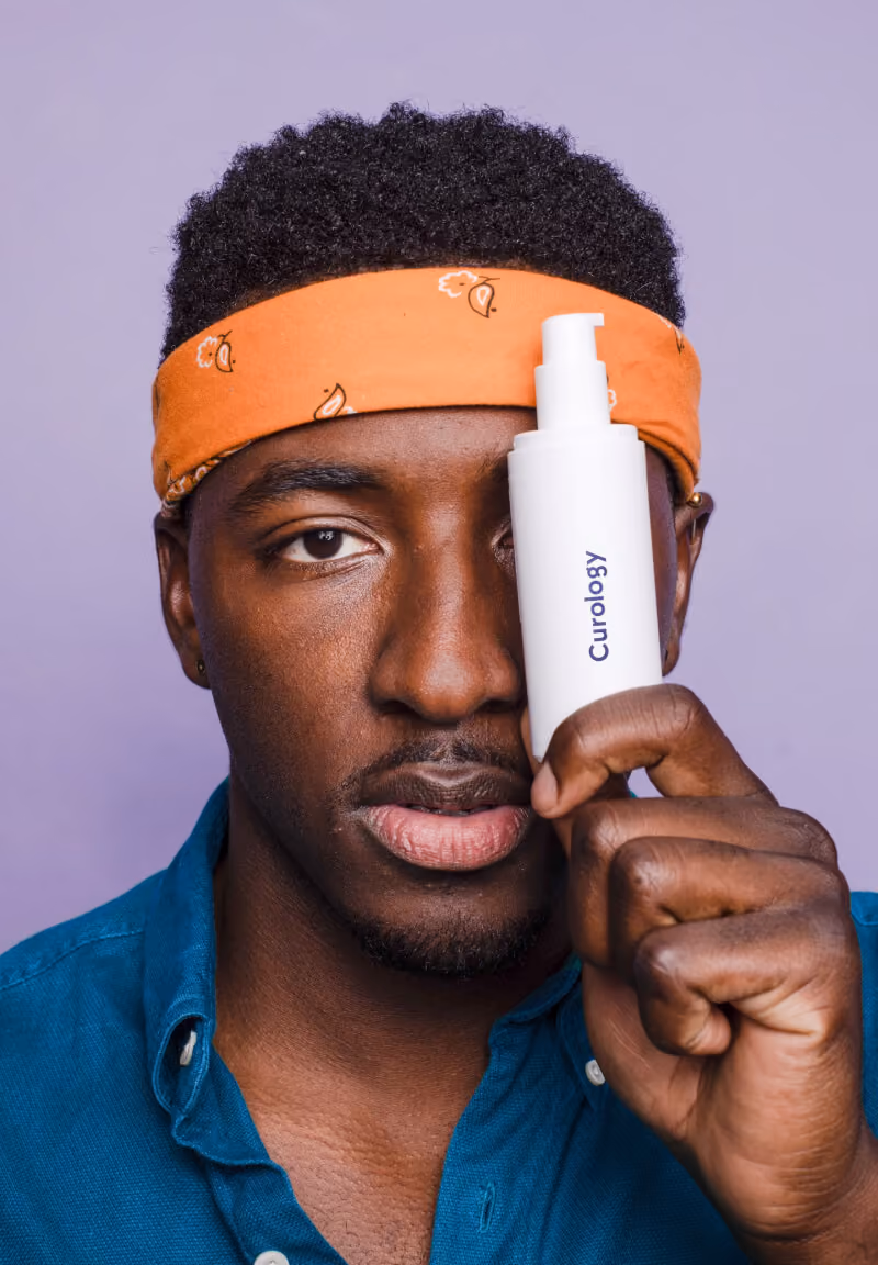 Man with orange headband holds a white Curology bottle near his right eye against a purple background.
