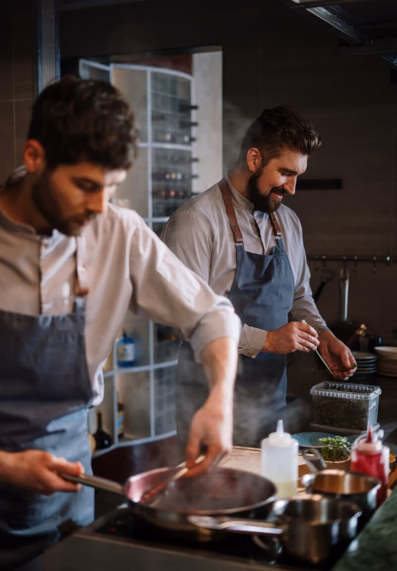 Two male chefs in aprons cooking in a kitchen, one stirring a pan and the other adding herbs.