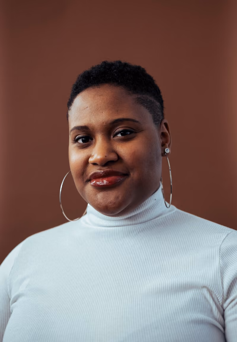Confident woman with short curly hair wearing a white turtleneck and large hoop earrings against brown background.