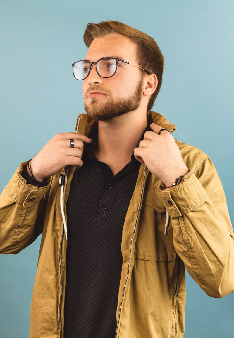 Bearded man with glasses adjusting the collar of his tan jacket against a light blue background.