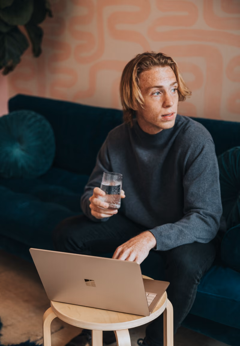 Young man sitting on a teal couch holding a glass of water with a laptop on a small round table.