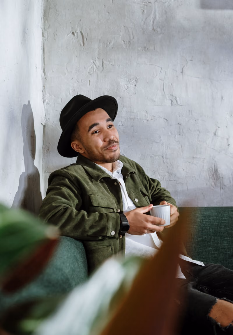 Man in a black hat and green jacket sitting on a couch holding a white cup, looking thoughtful.