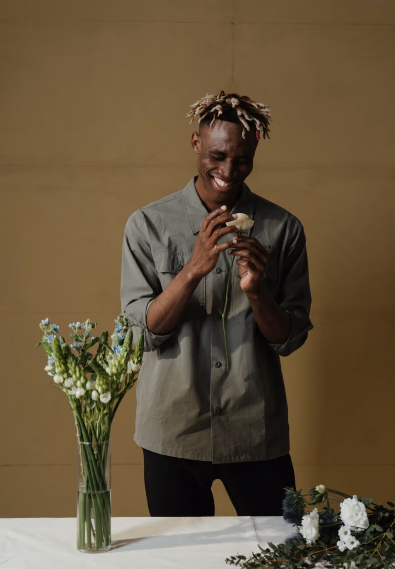 Man smiling while holding a white flower, standing behind a table with flowers and a vase.