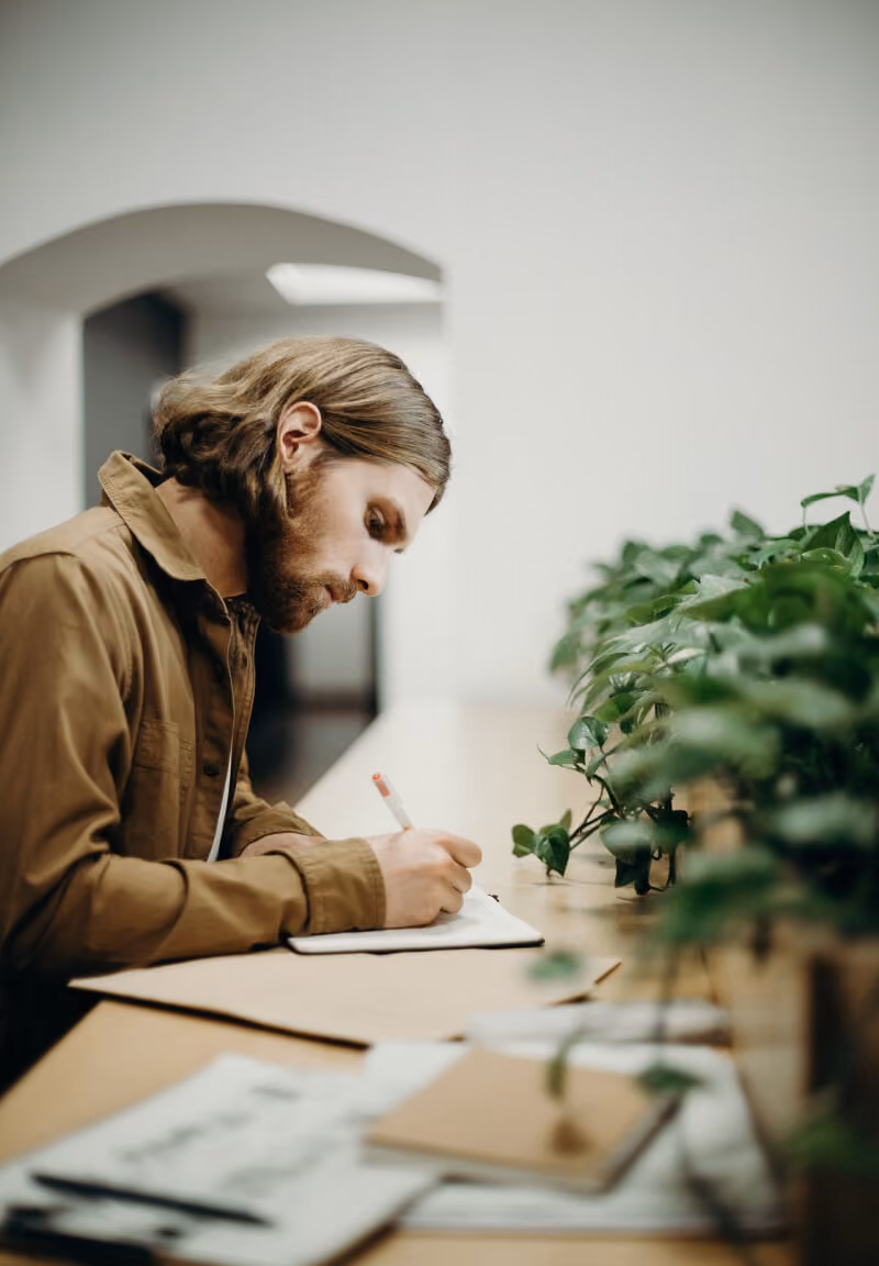 Bearded man with long hair focused on writing at a desk next to several green potted plants.