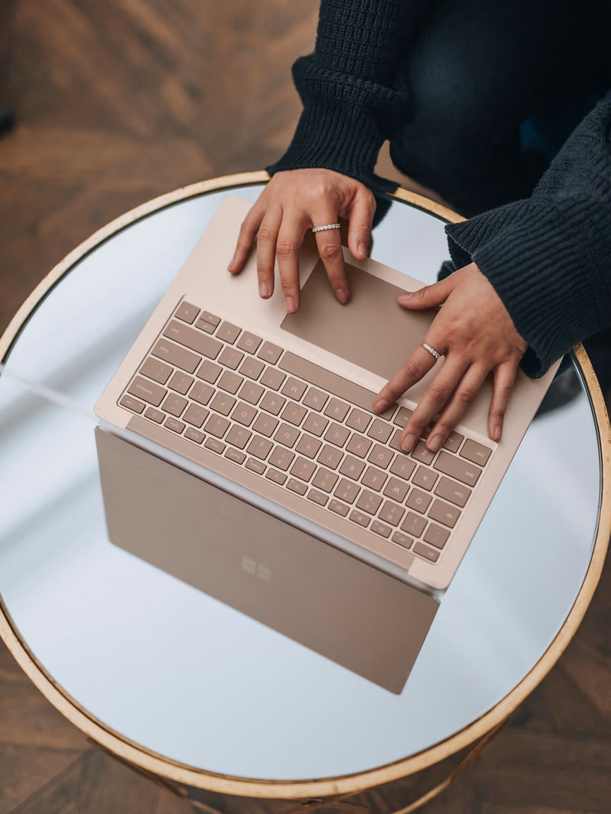 Person typing on a silver laptop keyboard on a round mirror table wearing dark sleeves and rings.
