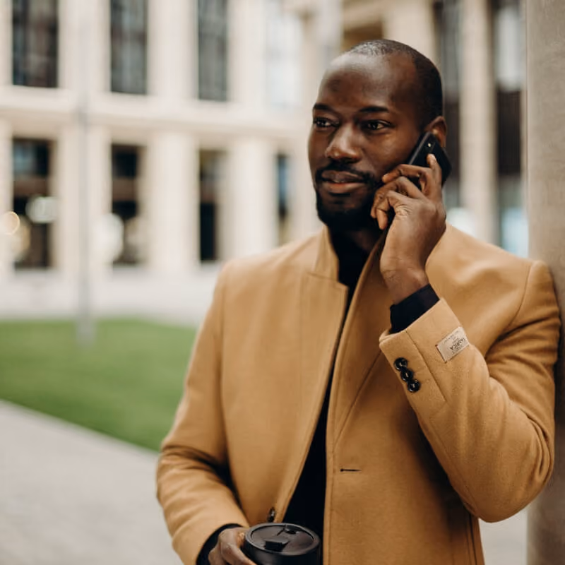 Man in tan coat talking on phone, holding coffee cup, standing outdoors by a column.