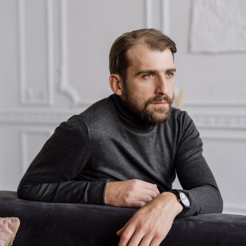 Bearded man in a dark turtleneck leaning on a dark sofa, looking contemplative to the side.