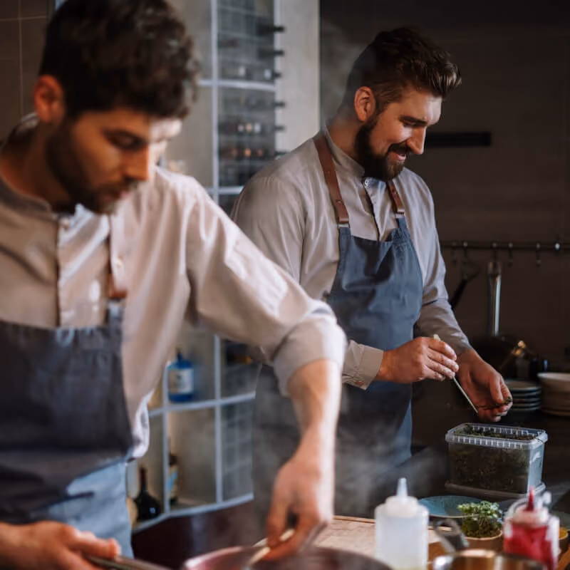 Two male chefs in aprons prepare food in a professional kitchen with steam rising nearby.