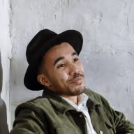 Man with mustache wearing a black hat and green jacket leaning against a white wall.