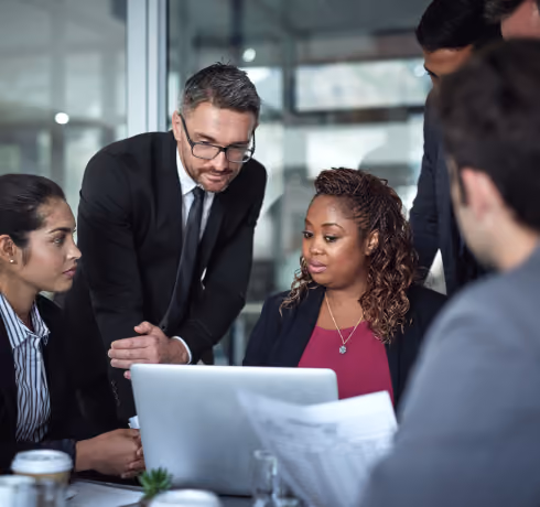 Five professionals in business attire gathered around a laptop, discussing work in an office.