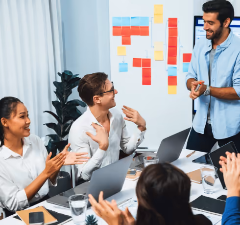 A diverse team applauds a presenter who stands by a whiteboard with sticky notes in a modern office.