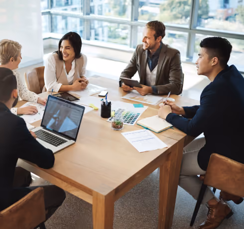 Five colleagues sit around a wooden table in a bright office, engaged in a discussion with laptops and papers.