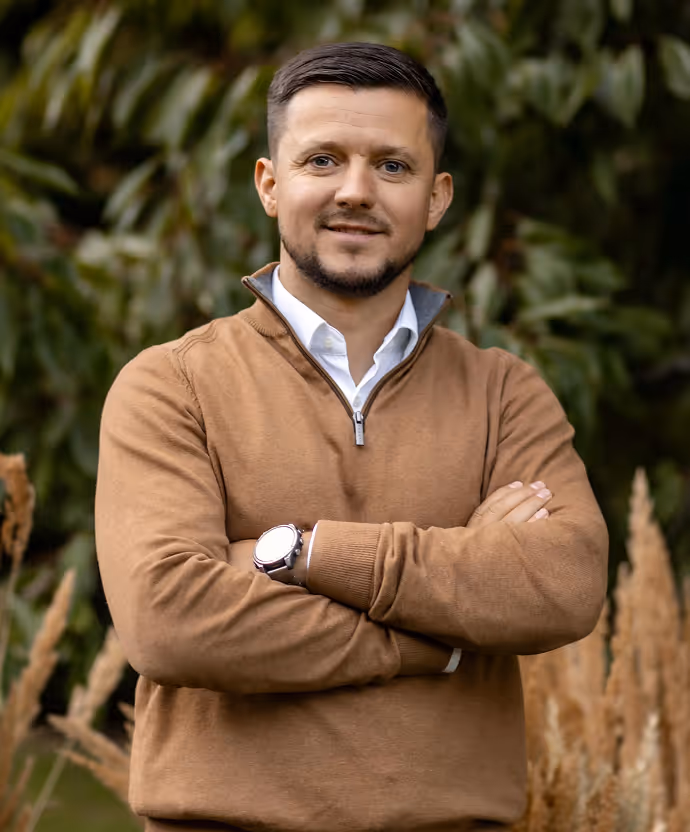 Man with short dark hair and beard wearing a brown quarter-zip sweater with a white collared shirt underneath, standing outdoors with arms crossed and greenery in the background.