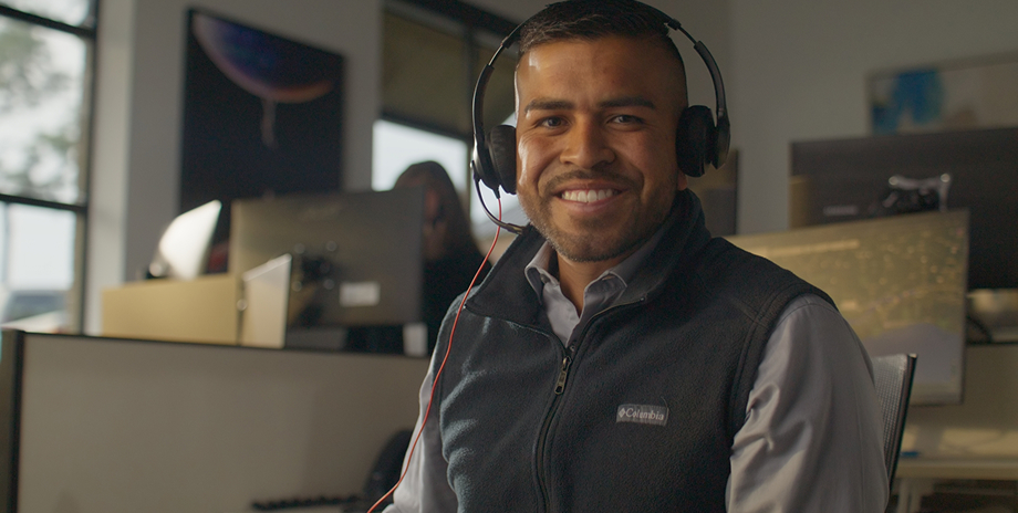 Smiling man wearing headphones and a Columbia vest sitting in an office with computer monitors in the background.