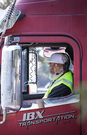 Truck driver wearing a white cap, headset, and yellow safety vest sitting in the red IBX Transportation truck cab.