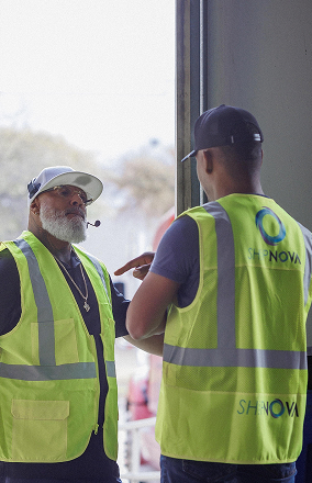 Two men wearing yellow reflective safety vests with SHINOVA logos, standing and talking near an open doorway.