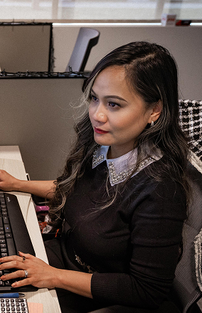 Woman with long dark hair wearing a black sweater with a decorative collar working at a computer in an office cubicle.