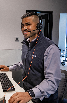 Smiling man wearing a headset and dark vest working at a computer in an office.