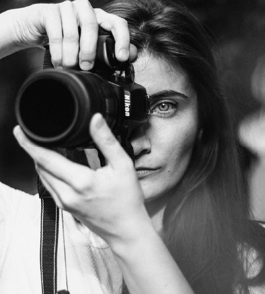 Black and white portrait of a woman taking a photo with a Nikon camera, focusing through the lens.