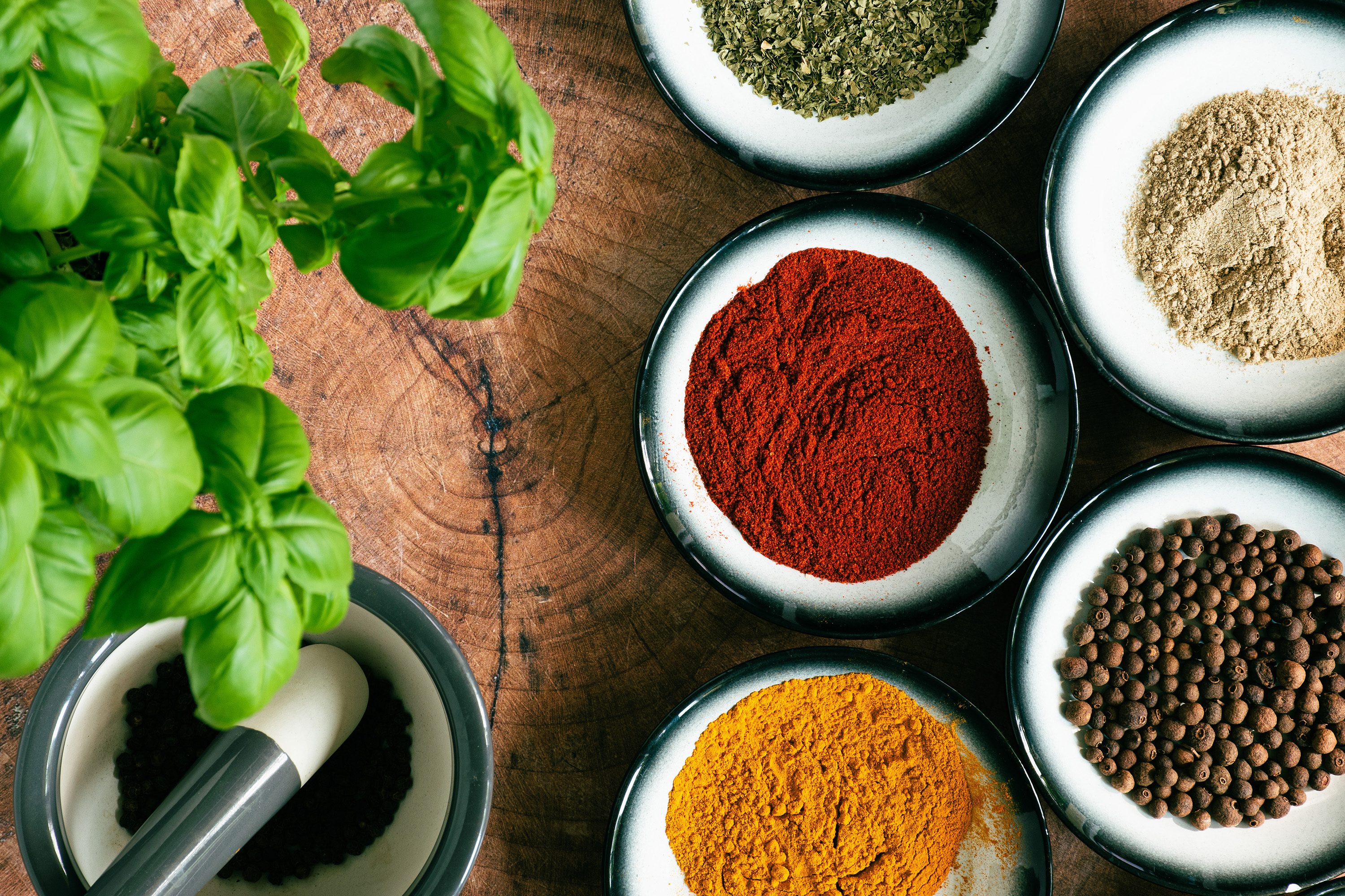 Różne przyprawy w miseczkach na drewnianym stole obok świeżej bazylii i moździerza z ziarnami pieprzu.