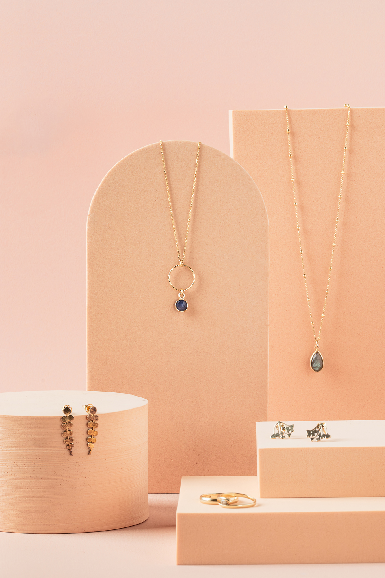 Delikatna złota biżuteria na pastelowym beżowym tle, w tym dwie naszyjniki, dwa pierścionki i dwie pary kolczyków.