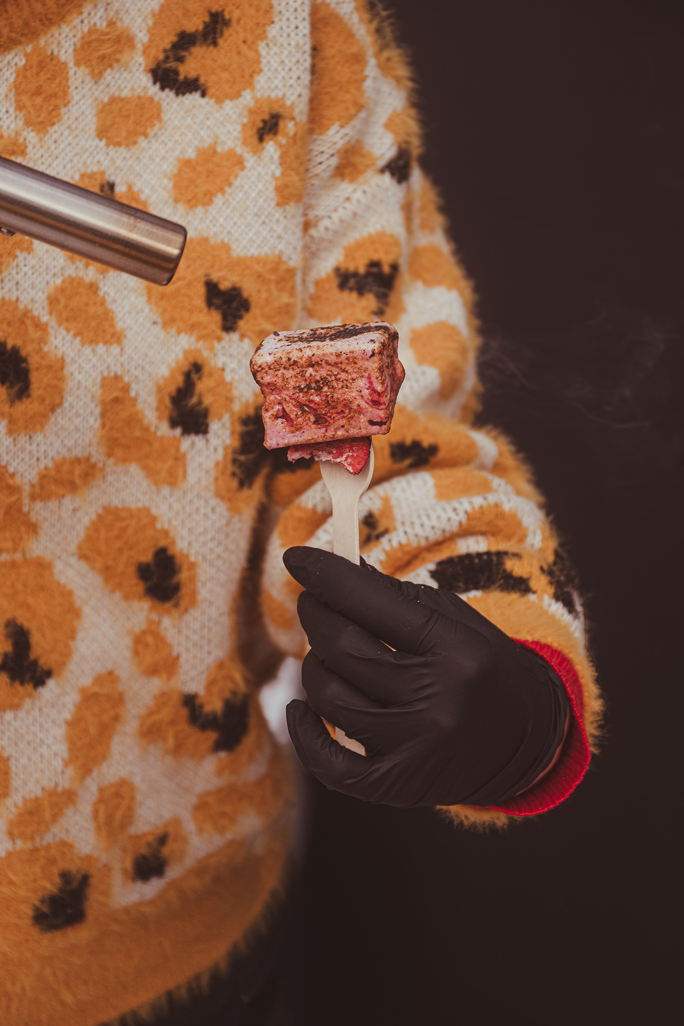 Osoba w czarnej rękawiczce trzyma drewniany widelec z zrumienionym piankowym deserem, obok dmuchawa gazowa, na tle pomarańczowo-białego swetra w wzór zwierzęcy.