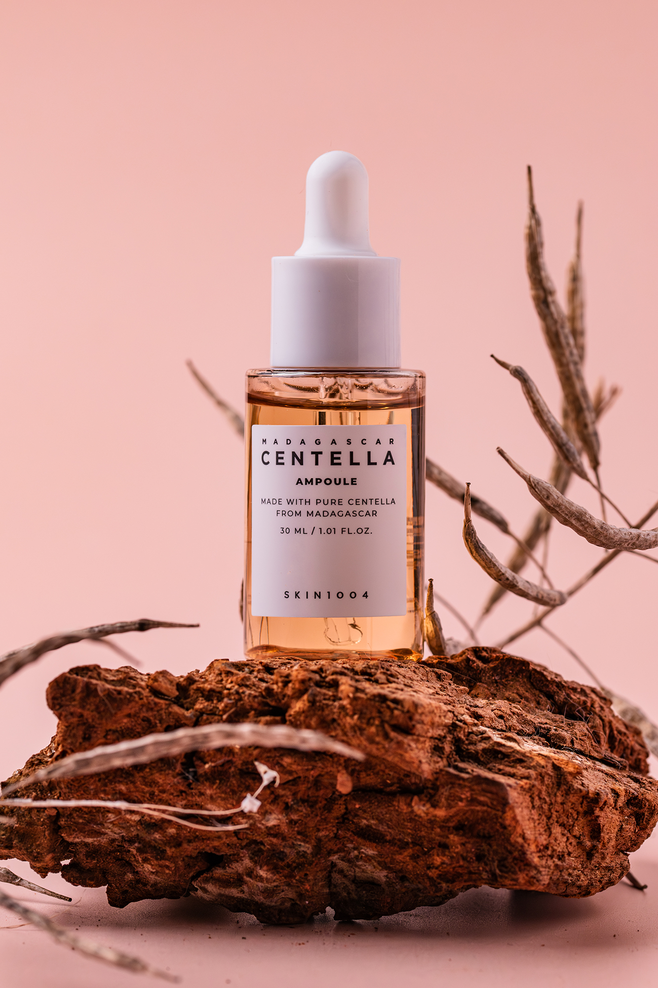 Butelka ampułki Madagascar Centella na kawałku drewna z suchymi roślinnymi gałązkami na różowym tle.