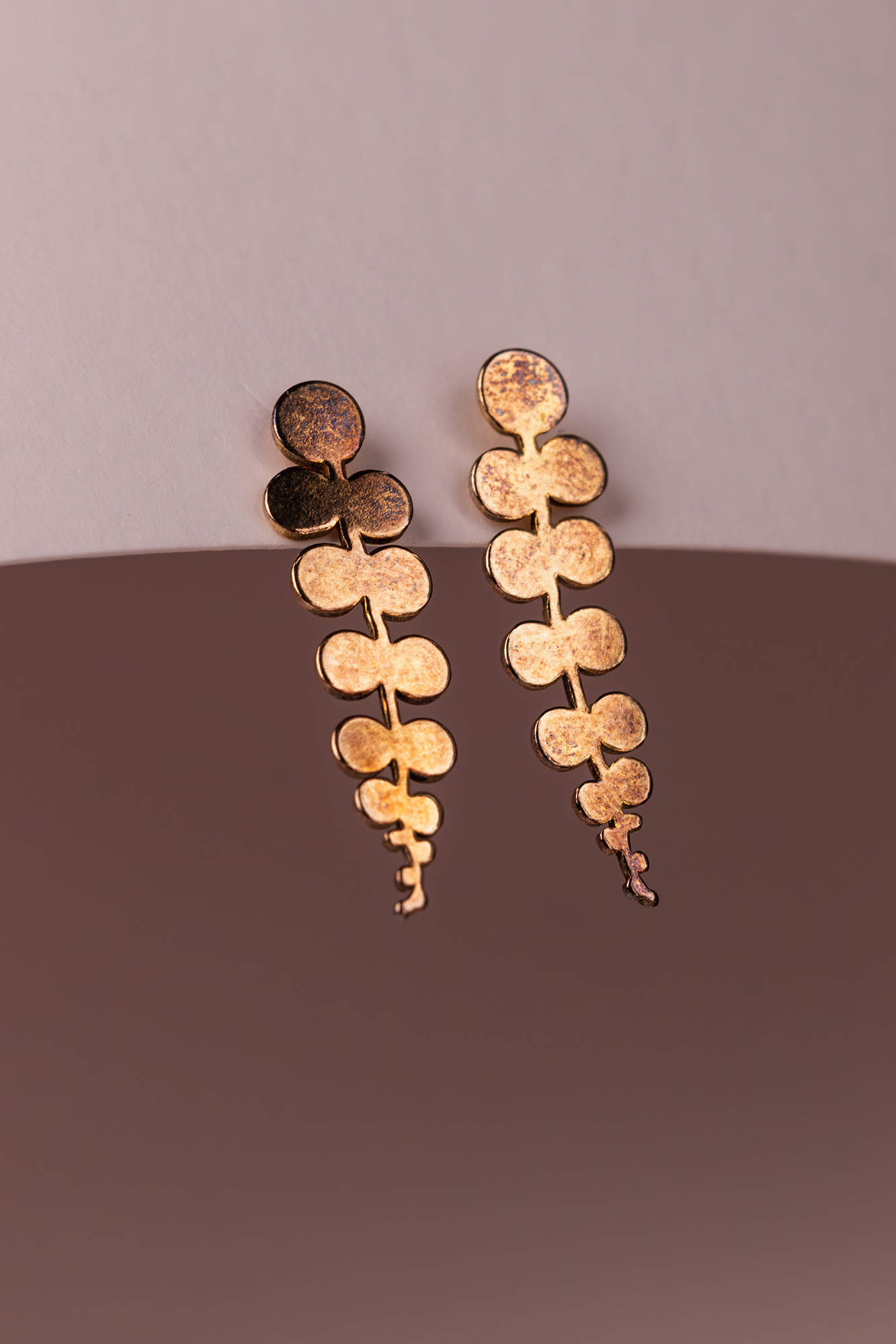 Para kolczyków ze złotym metalem w kształcie łączonych, zaokrąglonych elementów na pastelowym tle.