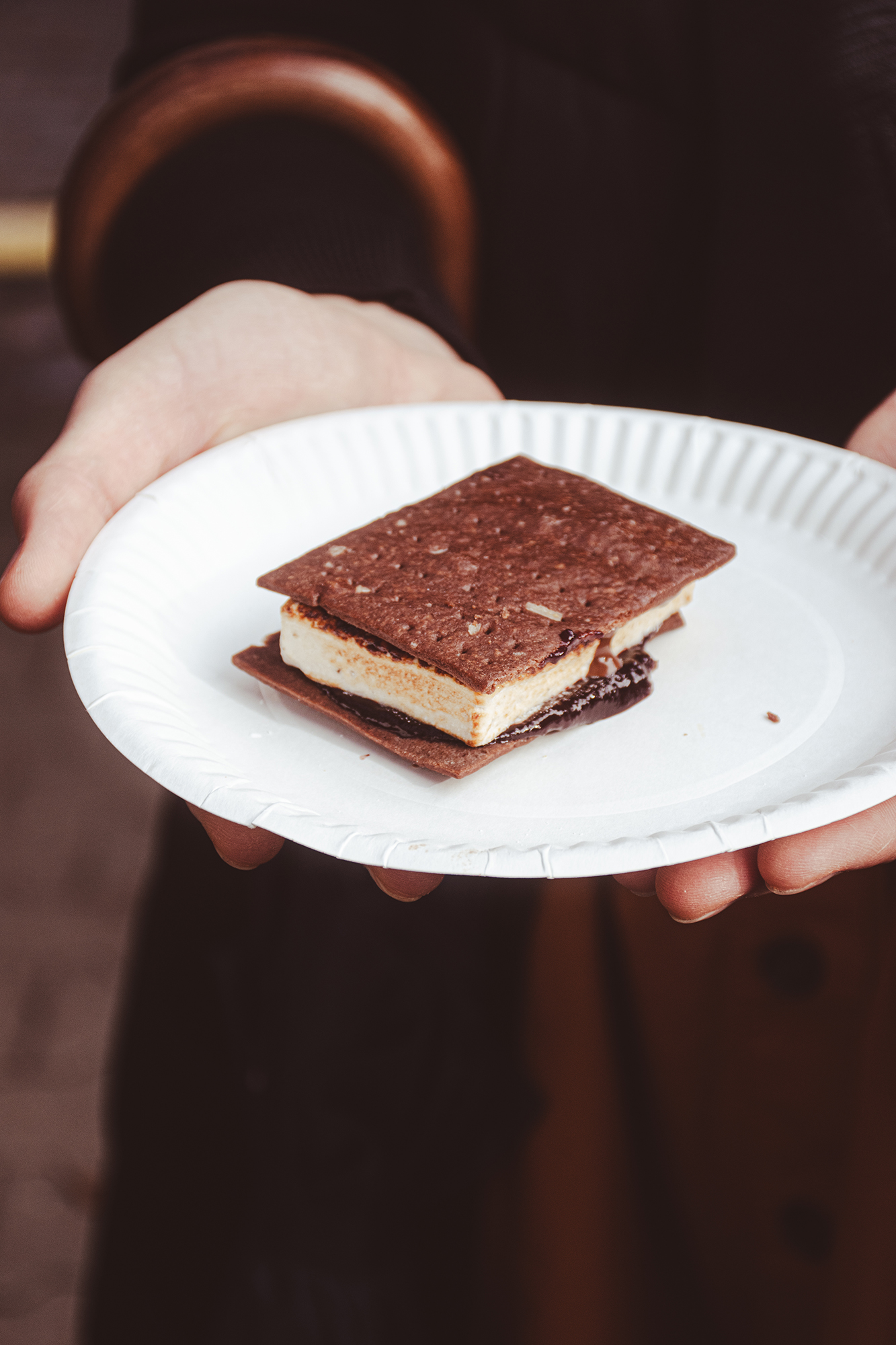 Ręce trzymające papierowy talerz z przygotowanym s'more z pianką między dwoma kawałkami czekoladowych herbatników.