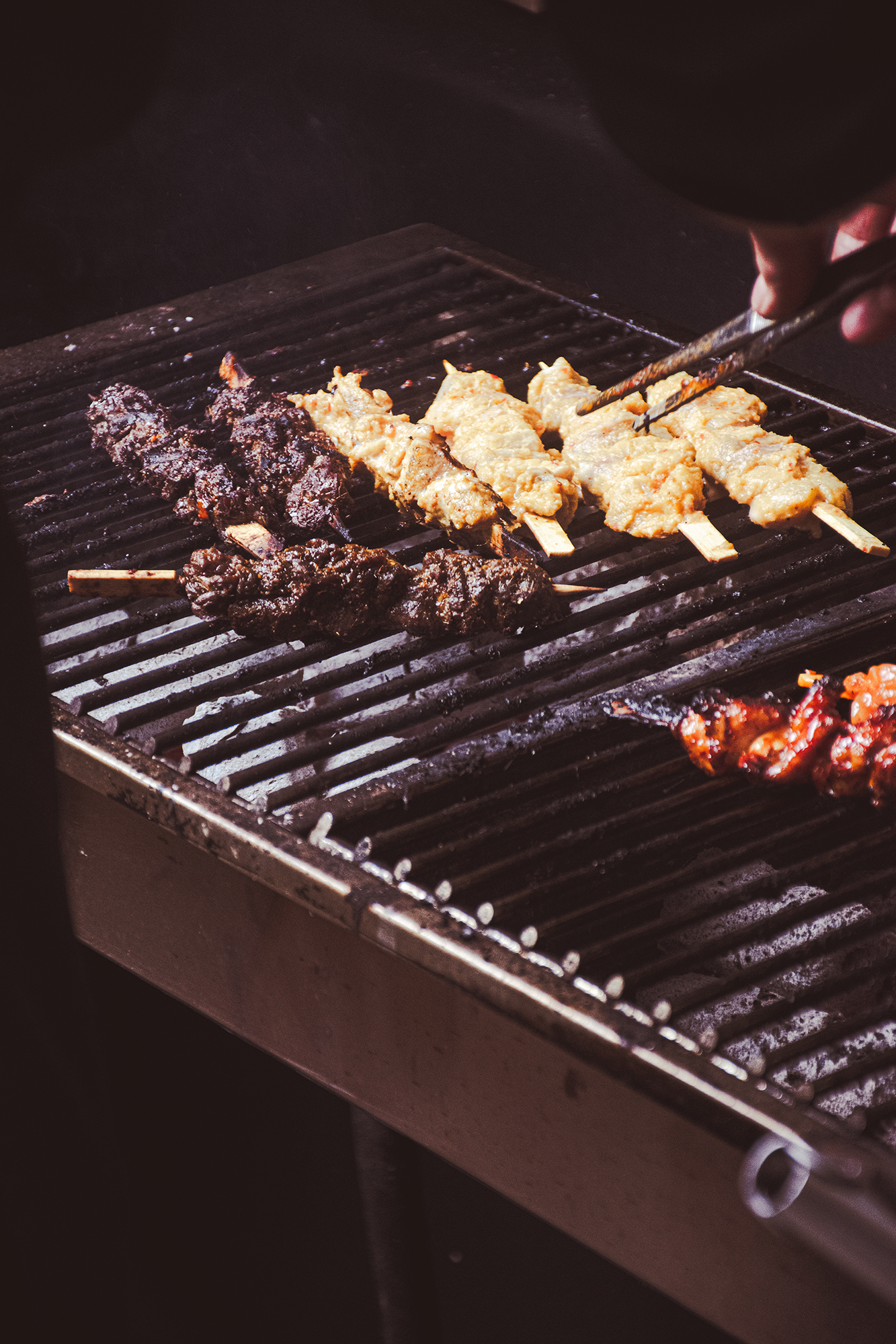 Różne mięsa na patyczkach grillowane na grillu węglowym, jedna ręka trzyma szczypce obracając kawałek mięsa.