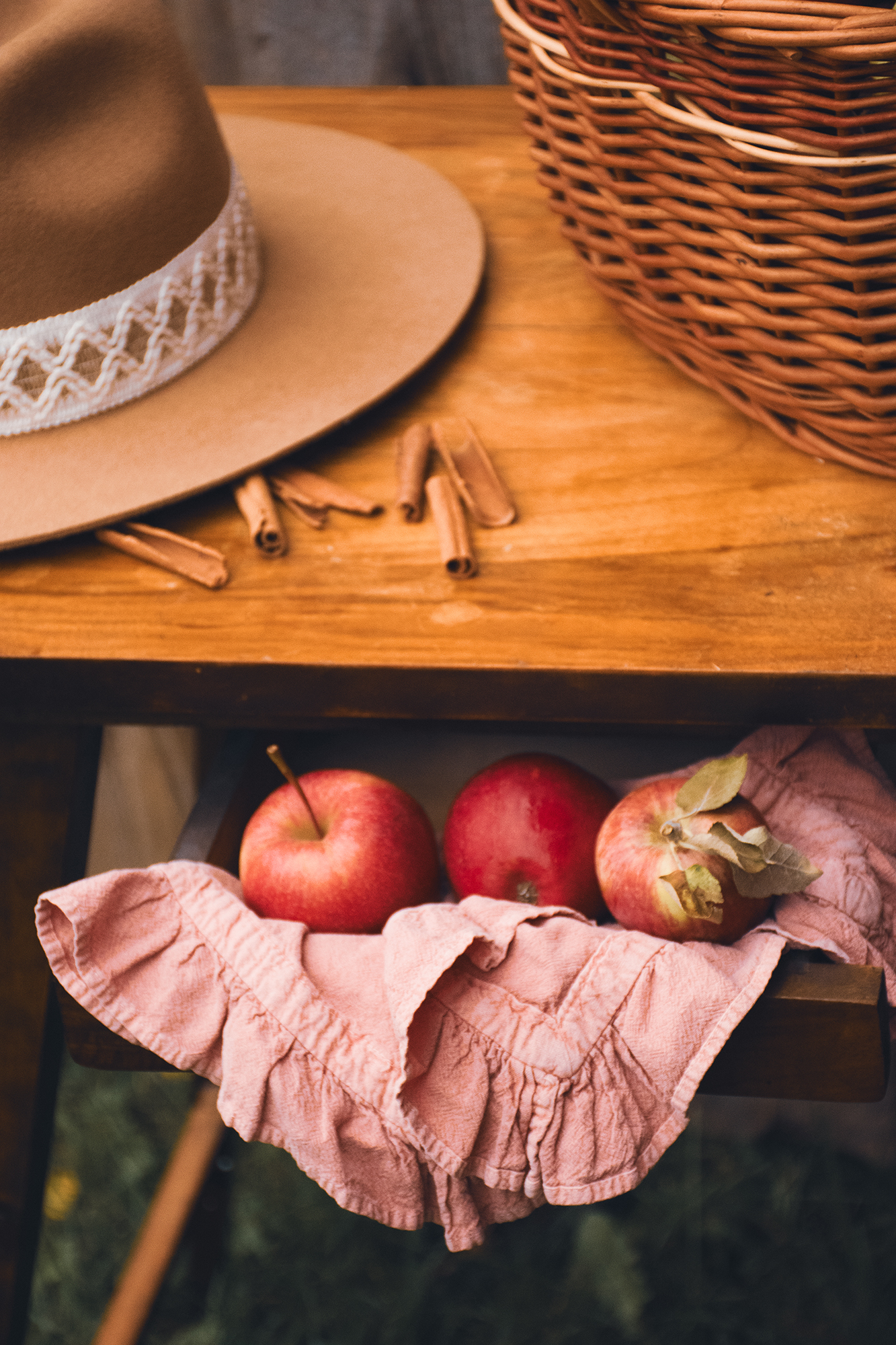 Trzy czerwone jabłka na różowej ściereczce w wysuniętej szufladzie drewnianego stolika, obok leży kapelusz i kilka lasek cynamonu.