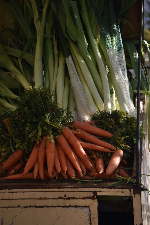 Pęk świeżych marchewek z zielonymi liśćmi ułożonych obok pęków długich zielonych porów na drewnianej powierzchni.