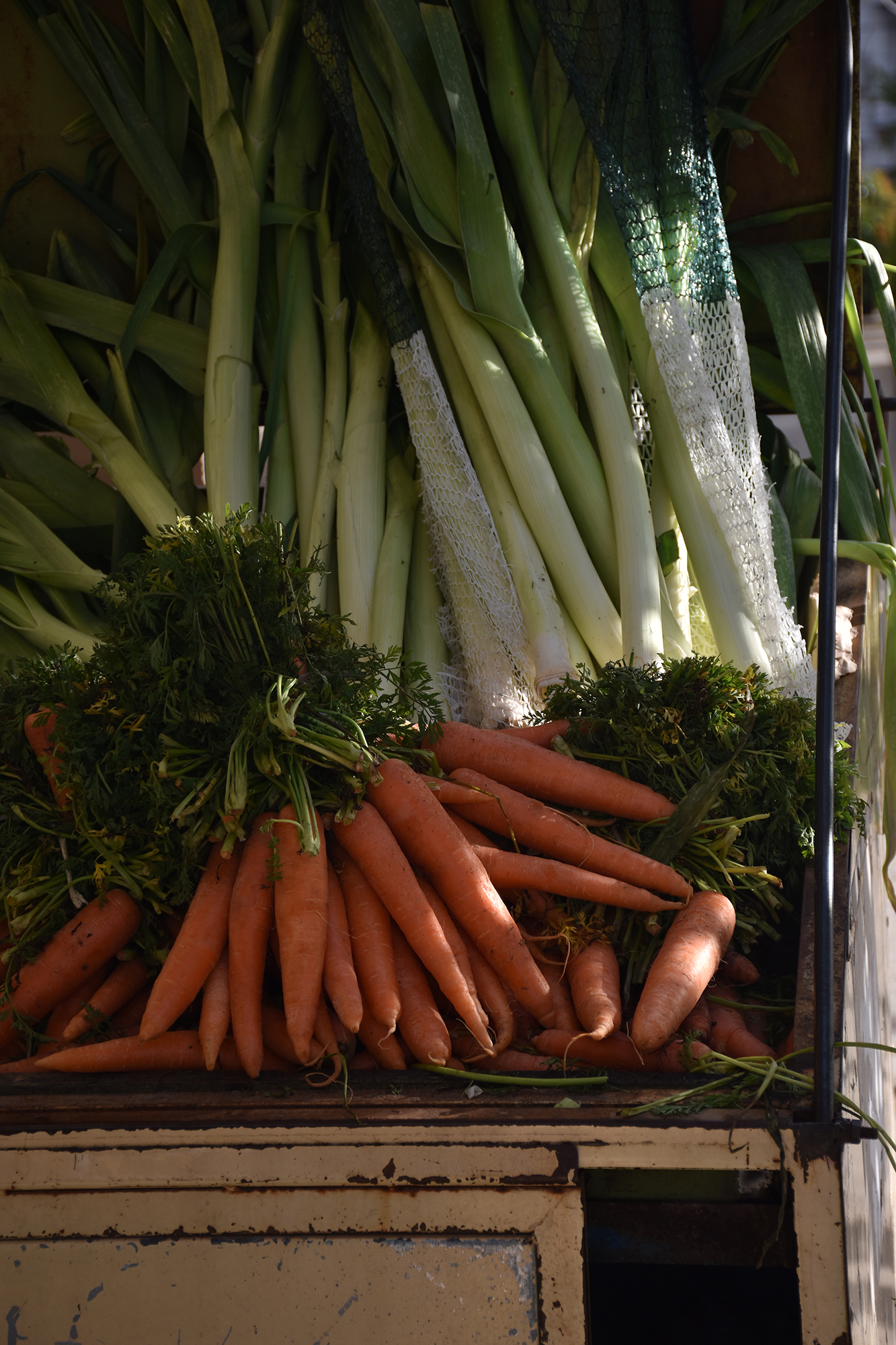 Pęk świeżych marchewek z zielonymi liśćmi ułożonych obok pęków długich zielonych porów na drewnianej powierzchni.