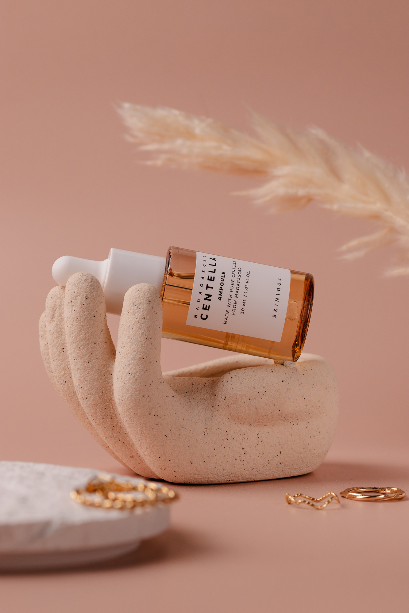 Butelkę ampułki z ekstraktem z centelli ustawiona na dekoracyjnej ceramicznej dłoni na różowym tle obok złotej biżuterii.