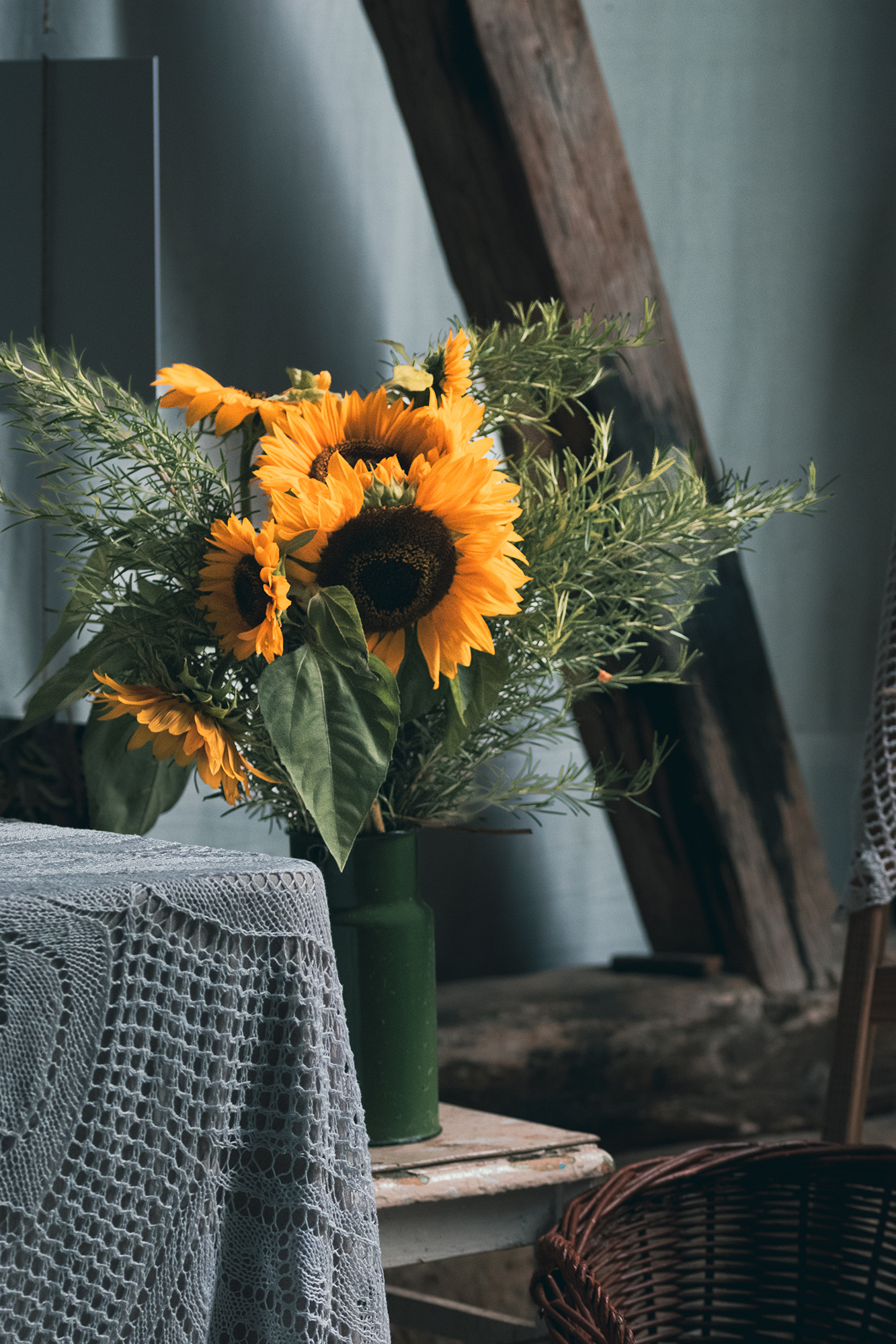 Bukiet słoneczników i zielonych liści w zielonym wazonie na drewnianym stole obok stołu z szydełkowym obrusem.