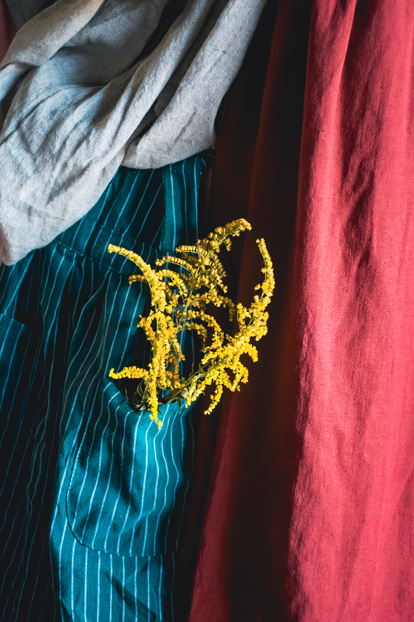 Żółte kwiaty w kieszeni pasiastego niebieskiego fartucha na tle czerwonej tkaniny i szarego materiału.