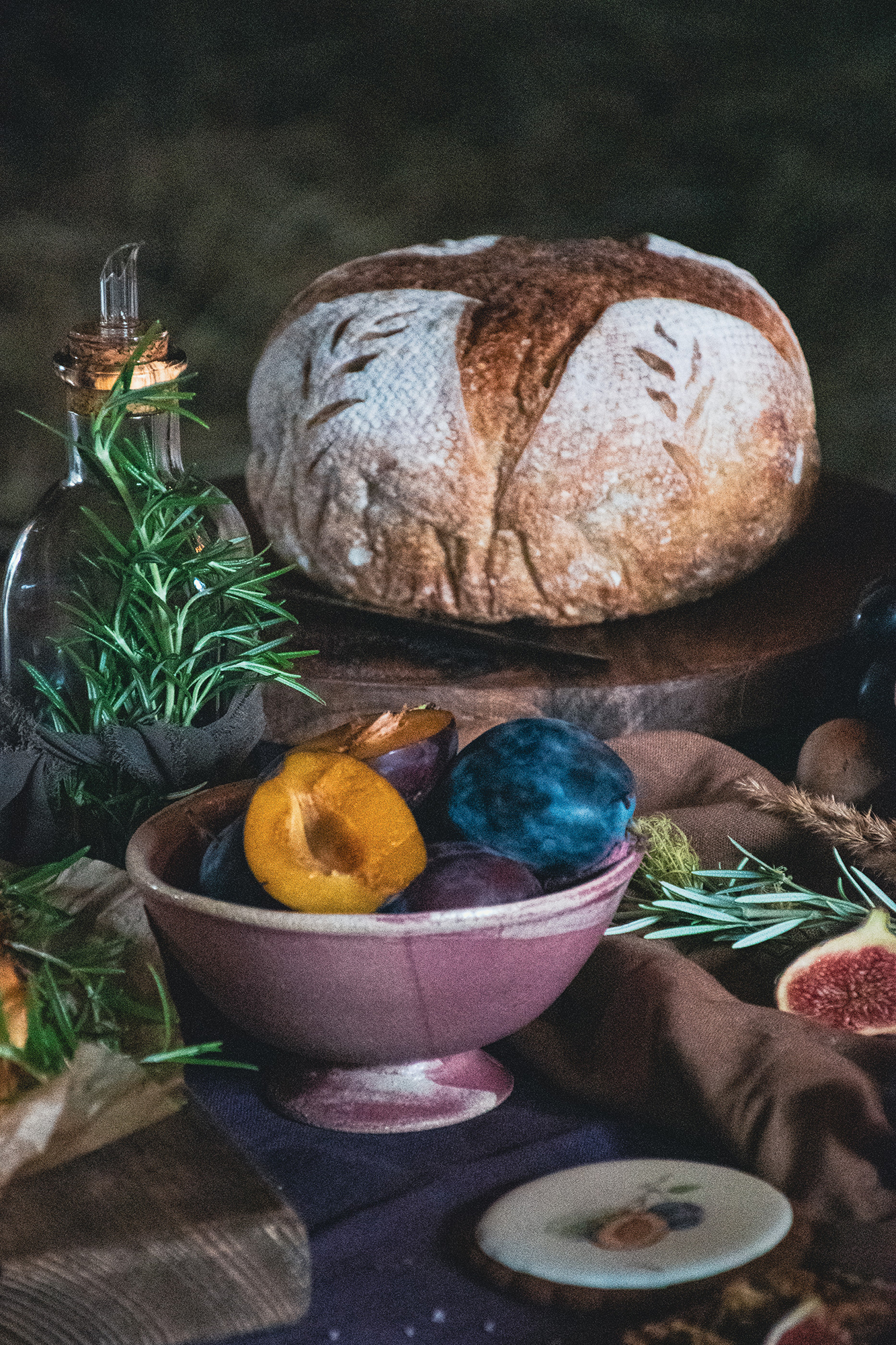 Bochenek chleba z dekoracyjnym wzorem, miska ze śliwkami i gałązki ziół na stole z ciemnym obrusie.