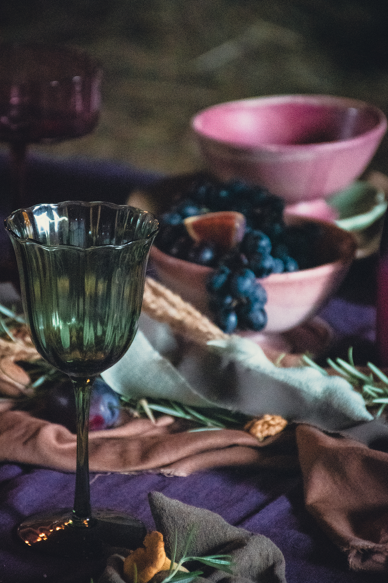 Zielony kielich na stole obok miski z ciemnymi winogronami i kawałkiem cytryny oraz dekoracji z ziół i tkanin.