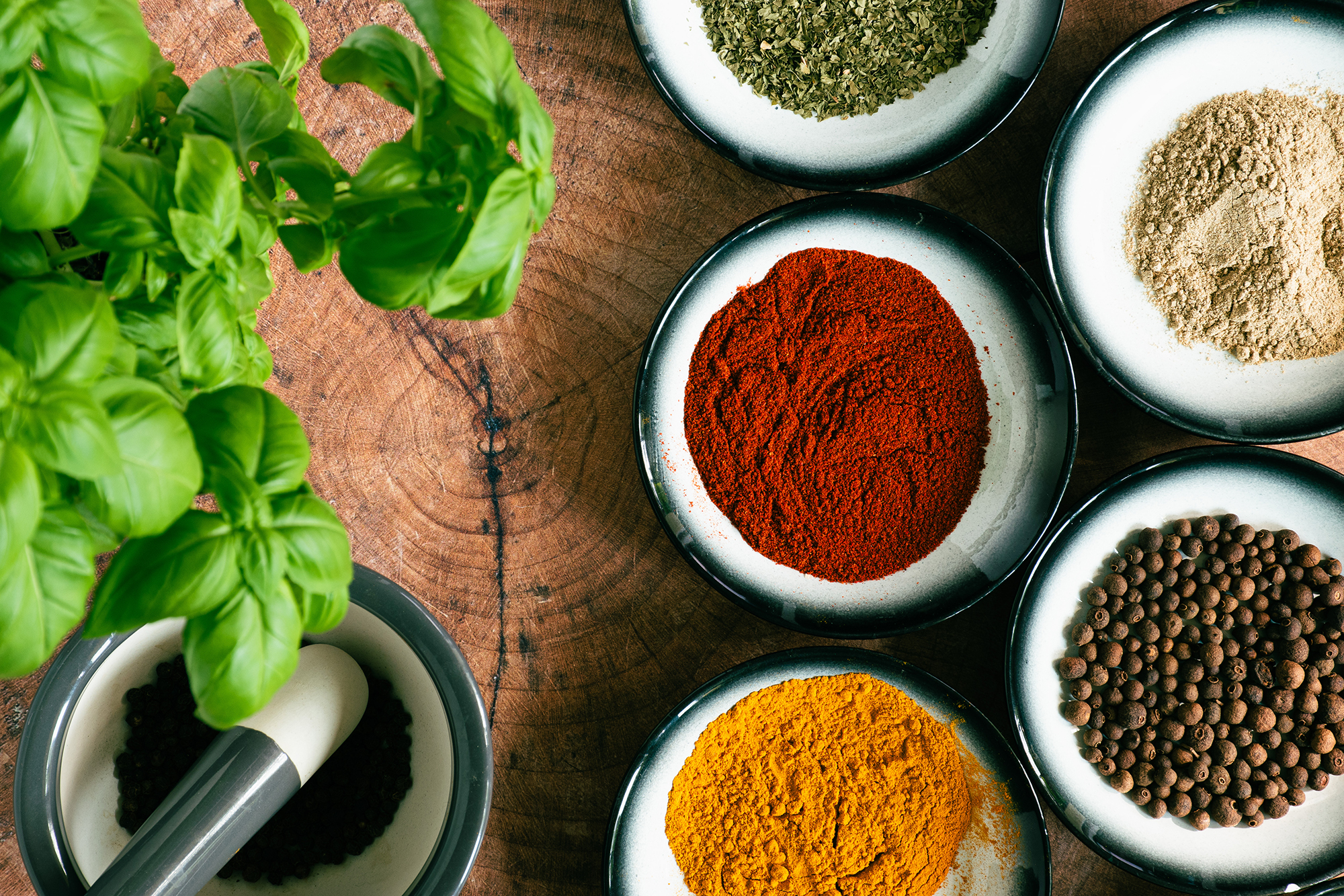 Różnokolorowe przyprawy w miseczkach i świeża bazylia na drewnianym blacie.