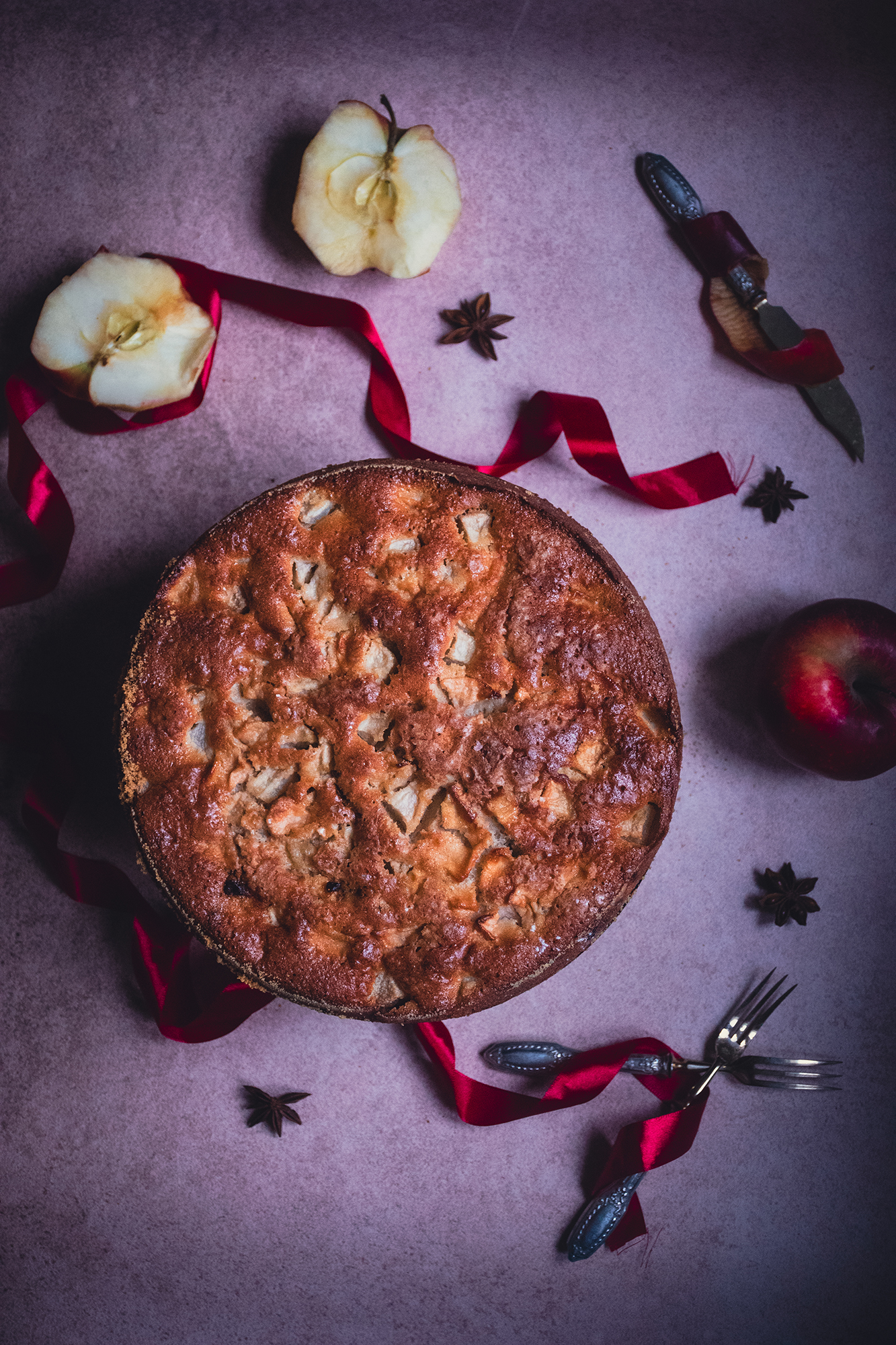 Jabłecznik na blacie z połówkami jabłek, czerwoną wstążką, anyżem, nożem i dwoma widelcami.